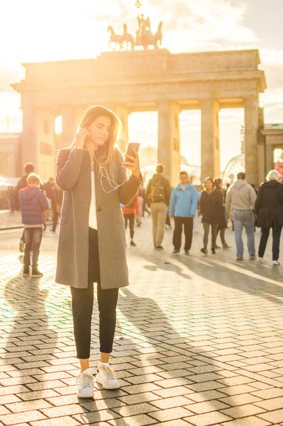 A fiatal nő teljes hossza hallgat zenét mobiltelefonon, miközben áll Brandenburg kapu Berlinben - Fotó, kép