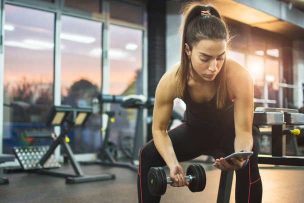 Sportliche Frau hält Handy und Hantel in der Pause im Fitnessstudio - Foto, Bild