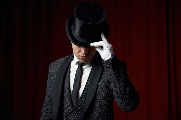 Portrait d'un jeune bel homme illusionniste portant un costume de tailleur festif touchant le bord du chapeau supérieur saluant avant une performance magique sur la scène du théâtre avec des draperies rouges - Photo, image