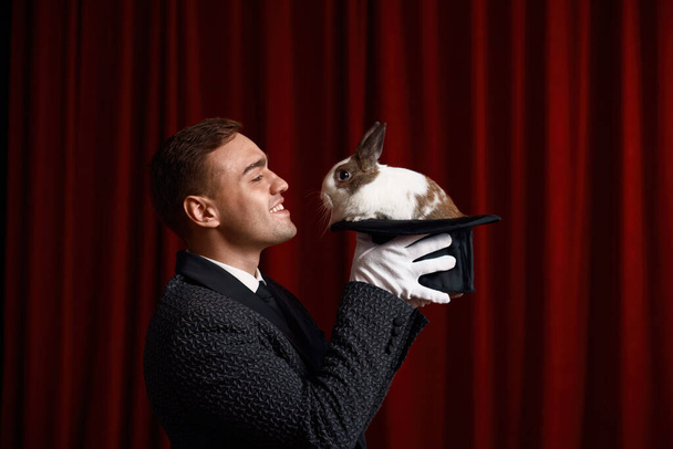 Tavşana bakan sihirbazın yan görünüş portresi şapkanın içinde belirdi. Tiyatro sahnesinde evcil hayvan gösterisiyle odaklanan profesyonel bir illüzyonist. Büyü ve hayal gücü kavramı - Fotoğraf, Görsel