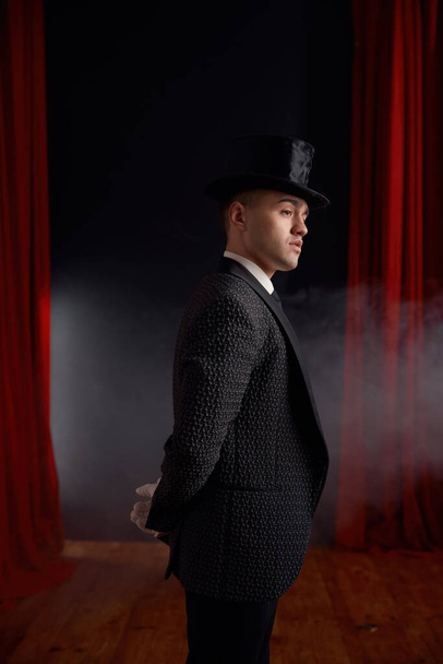Elegant man magician wearing elegant suit standing in smoke on stage looking at audience. Young showman entertainer against red drapery curtain side view - Photo, Image