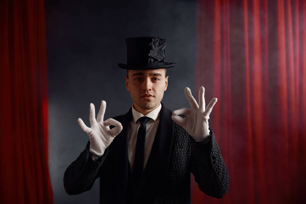 Retrato de showman en sombrero de copa y frac contra cortina roja sobre fondo en atmósfera misteriosa gesticulando con las manos usando guantes blancos realizando truco de magia - Foto, Imagen