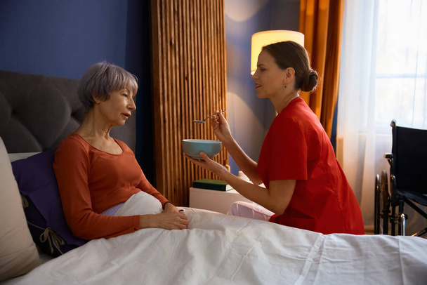Enfermera alimentando a anciana en el dormitorio de casa. Especialista en cuidadoras que brinda servicio de cuidado privado - Foto, Imagen