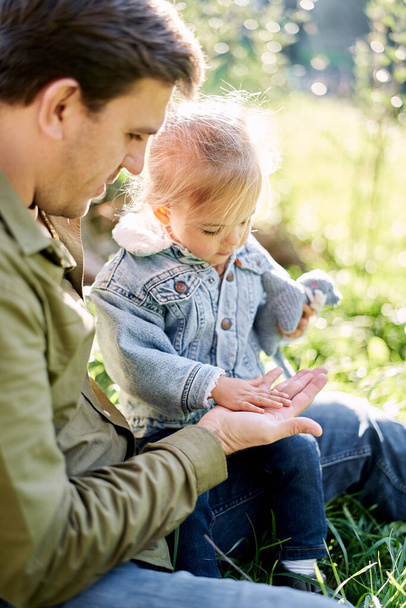 Das kleine Mädchen bedeckte die Handfläche ihres Vaters, während sie auf seinem Schoß auf dem Rasen saß. Hochwertiges Foto - Foto, Bild