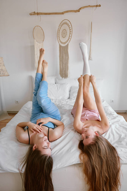 Two young beautiful girl friends wearing pajamas enjoying free time together lying on bed in bedchamber with raised up legs. Friendship and gossiping during slumber party celebration concept - Photo, Image