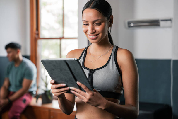 A woman is smiling while using a tablet computer, a portable communications device, at the gym. She holds it with her thumb, focusing on her thighs and chest during the event - Photo, Image