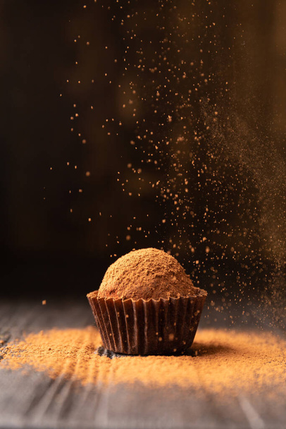 Kakaopulver wird bei schönem Licht auf eine Schokoladentrüffel gestreut. Eine Kugel Schokoladentrüffel wird in Großaufnahme mit Kakao auf dunklem Hintergrund bestreut. Konzeptfoto einer Gourmet-Trüffel. - Foto, Bild