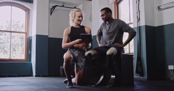 Fiatal afrikai férfi dupla amputált protézisekkel beszél a női személyi edző egy tabletta beltéri fitness tornaterem. Kiváló minőségű 4k felvételek. - Felvétel, videó