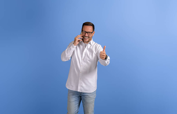 Felice giovane uomo d'affari mostrando pollici su segno e comunicare su smart phone su sfondo blu - Foto, immagini