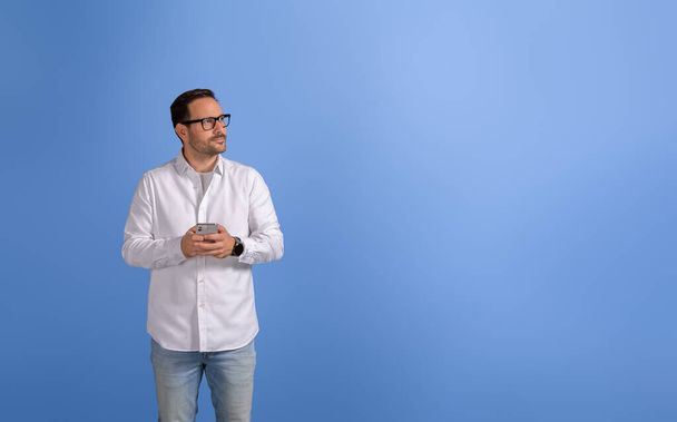 Задумчивый молодой бизнесмен с мобильным телефоном серьезно смотрит в сторону на изолированный синий фон - Фото, изображение