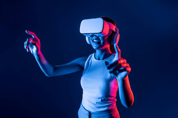 Mujer inteligente de pie en cyberpunk neón luz desgaste VR auriculares que conectan metaverso, futurista tecnología de la comunidad del ciberespacio. Mujer elegante uso dedo punto generado objeto virtual. Alucinaciones. - Foto, imagen