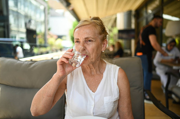 Happy senior woman perfect smile and teeth with glass of still water outdoors at summer sidewalk street cafe or restaurant terrace. Retired mature people holiday vacation, active lifestyle concept - Photo, Image