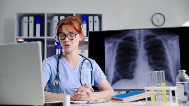 doctora con auriculares habla con el paciente a través de un enlace de video en el portátil y muestra una imagen de rayos X, durante una consulta médica en línea mientras está sentada en el consultorio médico - Imágenes, Vídeo