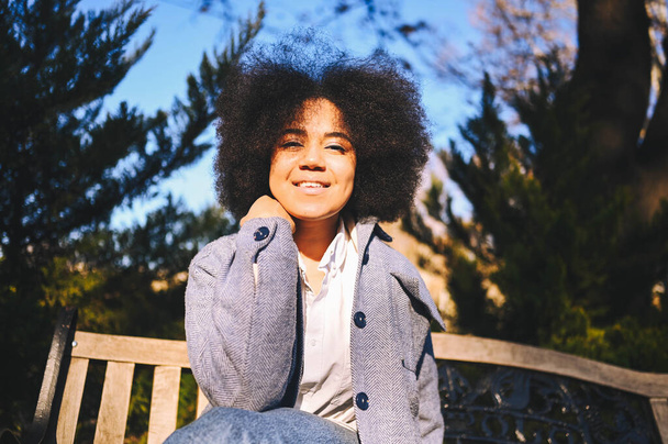 Close up Fashion street style portrait of attractive young natural beauty African American woman with afro hair in blue coat posing walking outdoors in sunny day. Happy lady with perfect teeth smile - Fotografie, Obrázek