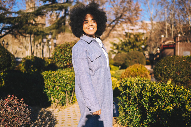 Close up Fashion street style portrait of attractive young natural beauty African American woman with afro hair in blue coat posing walking outdoors in sunny day. Happy lady with perfect teeth smile - Valokuva, kuva