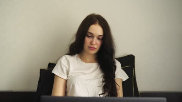 Keskittynyt nuori nainen katselee kannettavaa tietokonetta kirjoittaessaan, istuu sängyssä makuuhuoneessa. Hidasta vauhtia. Työskentely kotona käsite. Pääkonttori - Materiaali, video