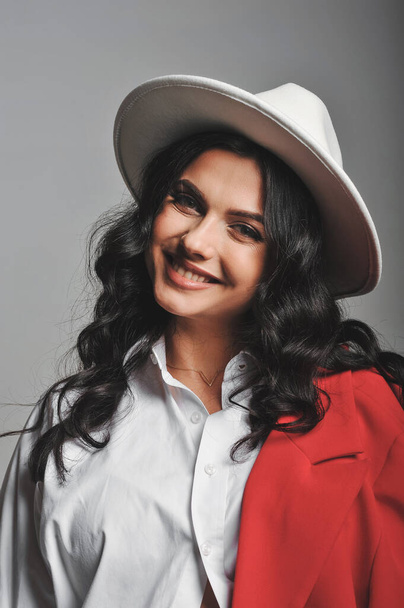 Ritratto di donna bruna sorridente in cappello bianco, camicia e giacca rossa in studio. Buono per carta da parati o rivista di stampa. Bellezza e moda - Foto, immagini