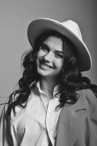 Ασπρόμαυρο πορτραίτο μιας χαμογελαστής μελαχρινής γυναίκας με λευκό καπέλο, πουκάμισο και κόκκινο σακάκι στο στούντιο. Καλό για ταπετσαρία ή περιοδικό εκτύπωσης. Ομορφιά και μόδα - Φωτογραφία, εικόνα