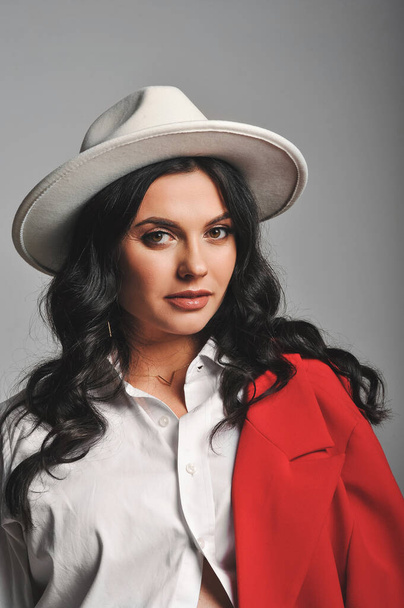 Πορτρέτο της όμορφης μελαχρινής γυναίκας σε λευκό καπέλο, πουκάμισο και κόκκινο σακάκι στο στούντιο. Καλό για ταπετσαρία ή περιοδικό εκτύπωσης. Ομορφιά και μόδα - Φωτογραφία, εικόνα