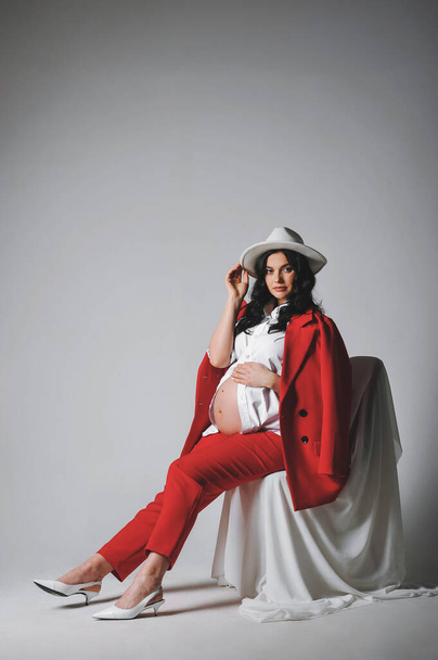 Εκπληκτική μελαχρινή έγκυος γυναίκα με λευκό καπέλο, πουκάμισο και κόκκινο κοστούμι που ποζάρει σε ένα κυκλόραμα. Αντιγραφή χώρου για κείμενο. Καλό για ταπετσαρία ή περιοδικό εκτύπωσης - Φωτογραφία, εικόνα