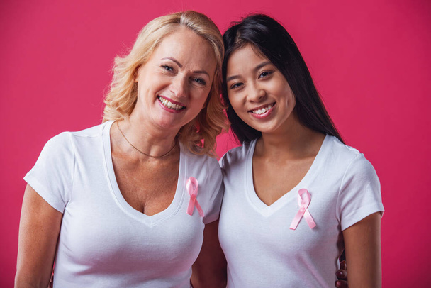 Frauen gegen Brustkrebs. schöne reife und asiatische Frauen mit rosa Schleifen auf der Brust schauen in die Kamera und lächeln auf rotem Hintergrund - Foto, Bild