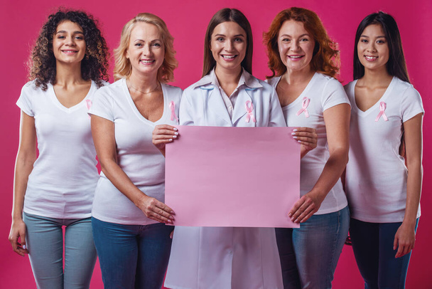 Mulheres contra o cancro da mama. Mulheres bonitas de diferentes idades e nacionalidades com fitas cor-de-rosa no peito estão segurando um pôster rosa e sorrindo, em fundo vermelho
 - Foto, Imagem