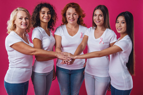 Women against breast cancer. Beautiful women of different ages and nationalities with pink ribbons on their chests are holding hands together and smiling, on red background - Photo, Image