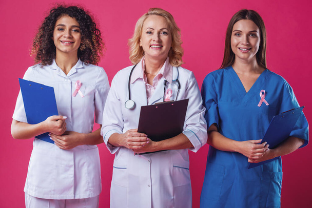 Жінки проти раку молочної залози. Красиві молоді жінки-лікарі з рожевими стрічками на грудях тримають папки, дивлячись на камеру і посміхаючись, на червоному тлі
 - Фото, зображення