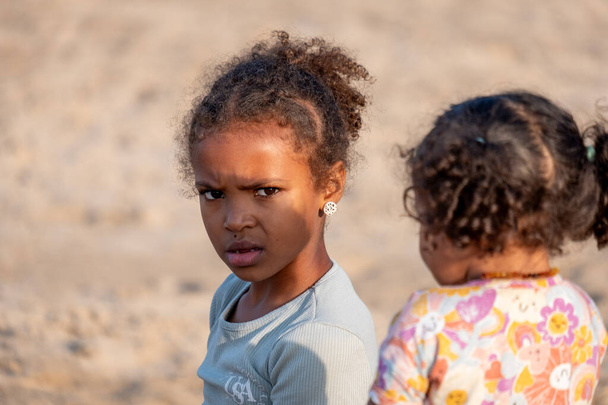 A pensive mixed race older girl looks back over her shoulder with a subtle frown, as a younger child faces away, depicting a moment of childhood introspection. High quality photo - Photo, Image