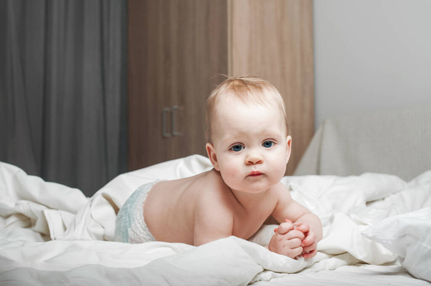Портрет красивого ребенка 11 месяцев в подгузнике на кровати крупным планом. Skincare, медицинская концепция для детей. Зубы, руки на рот малыша. - Фото, изображение