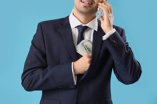Young businessman with money talking by mobile phone on blue background, closeup - Photo, Image