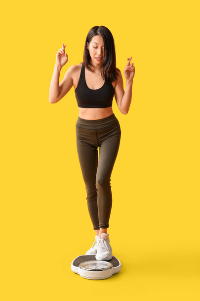 Beautiful young Asian woman with crossed fingers and scales on yellow background. Weight loss concept - Foto, immagini