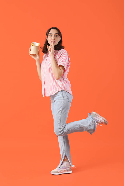 Giovane donna in possesso di patatine fritte e mostrando silenzio gesto su sfondo arancione - Foto, immagini