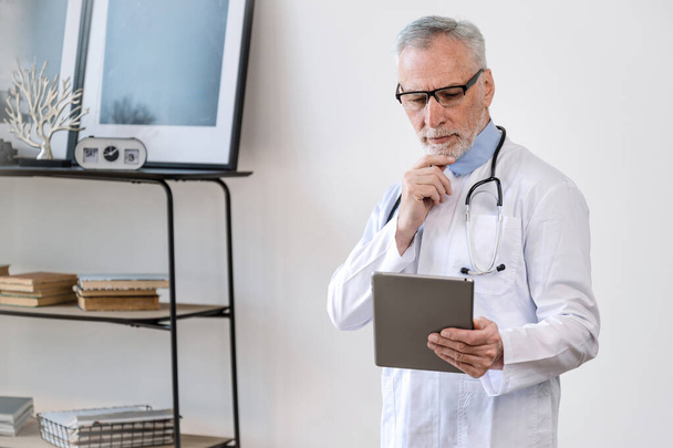Serious thoughtful professional doctor with stethoscope working in private clinic, holding modern touch screen gadget and analyzing patient diagnosis. Concept of telemedicine. - Photo, Image