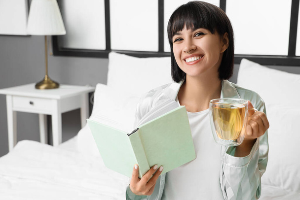 Giovane donna con tazza di tè lettura libro sul letto a casa - Foto, immagini