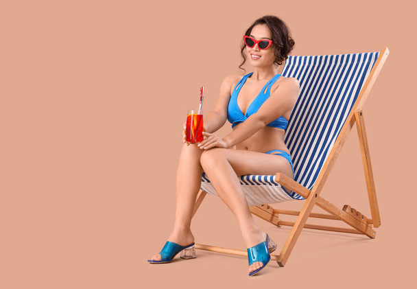 Sexy young woman in swimsuit and with cocktail relaxing on deck chair against beige background - Photo, Image