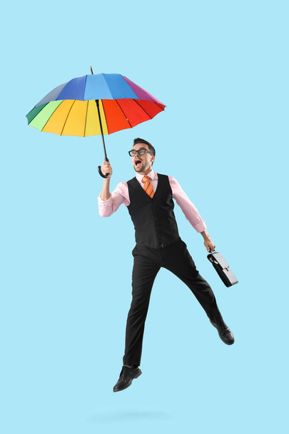 Αστείο σοκαρισμένος επιχειρηματίας με ομπρέλα και χαρτοφύλακα άλμα σε μπλε φόντο - Φωτογραφία, εικόνα