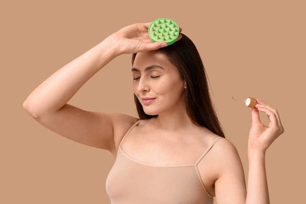 Mooie jonge vrouw met hoofdhuid massager en cosmetische olie op bruine achtergrond - Foto, afbeelding
