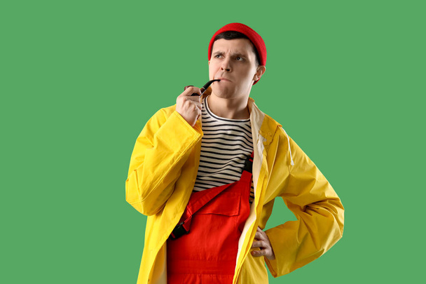 Thoughtful young sailor in raincoat with smoking pipe on green background - Фото, изображение