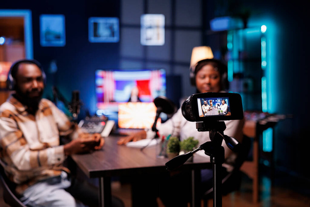 During live broadcast, podcast host records guest sharing engaging story with audience by mounting professional device on a tripod. Camera taking video of bloggers delivering facts in home studio. - Photo, Image