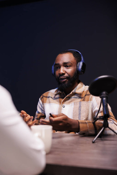 Czarny gospodarz przesłuchuje osobę w domu, używając nowoczesnego mikrofonu i bezprzewodowych słuchawek do swojego internetowego talk show. Afrykański Amerykanin rozmawiający z osobą nagrywającą odcinek podcastu. - Zdjęcie, obraz