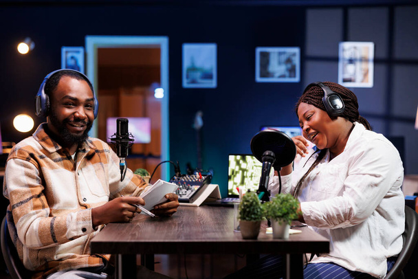 Συνέντευξη καταγραφή διασκεδαστικό συζήτηση με κωμικό κατά τη διάρκεια podcast γυρίστηκε στο στούντιο διαμέρισμα. Μαύροι άνδρες και γυναίκες vloggers χρησιμοποιώντας βιντεοκάμερα για την παραγωγή κωμικών talk show από το σπίτι - Φωτογραφία, εικόνα