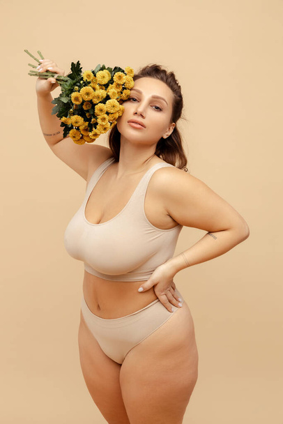 Retrato de mujer atractiva cuerpo positivo usando lencería sexy con estilo, sosteniendo ramo de crisantemo amarillo, de pie aislado sobre fondo beige. Concepto del Día Internacional de la Mujer - Foto, Imagen