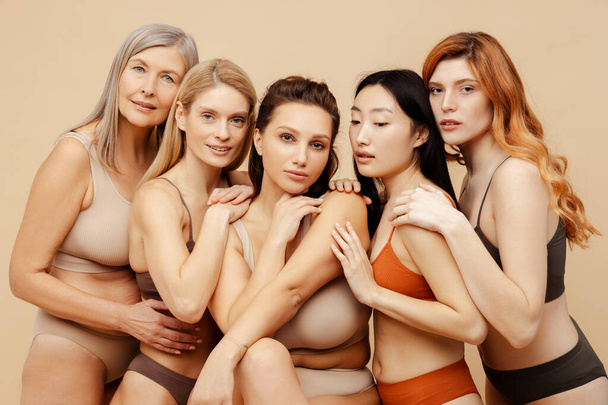 Group of multiethnic, diverse, attractive women wearing sexy lingerie, looking at camera, hugging. Beautiful, sensual fashion models posing isolated on beige background. International woman`s day - Photo, Image