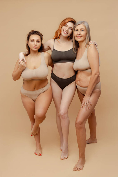 Cheerful, attractive, diverse women wearing stylish underwear, hugging and looking at camera. Beautiful fashionistas posing on beige background, in studio. Concept of natural beauty - Photo, Image