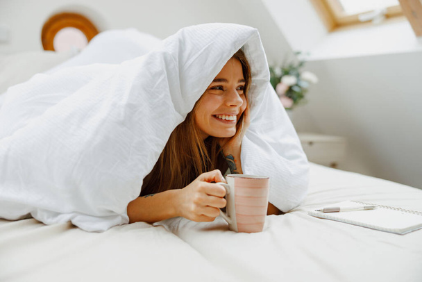 Γυναίκα χαμογελά στο κρεβάτι, κρατώντας φλιτζάνι καφέ με μανίκι, χαρούμενος - Φωτογραφία, εικόνα