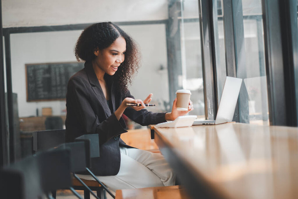 Una donna d'affari attenta con una tazza di caffè in una mano e uno stilo nell'altra lavora diligentemente sul suo computer portatile nell'atmosfera vibrante di un caffè - Foto, immagini