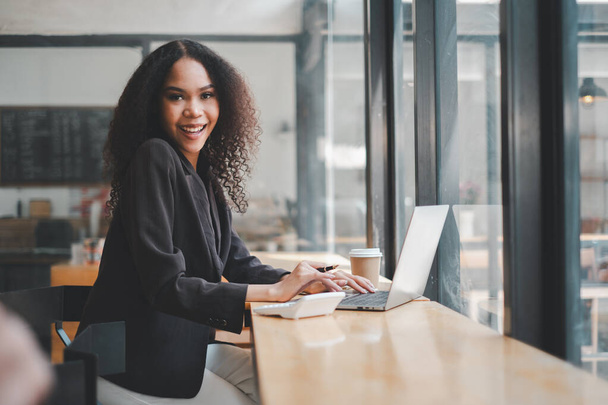 Avec un sourire éclatant, une jeune femme d'affaires est absorbée dans son travail sur un ordinateur portable dans un café, son énergie positive correspondant à l'ambiance animée autour d'elle. - Photo, image