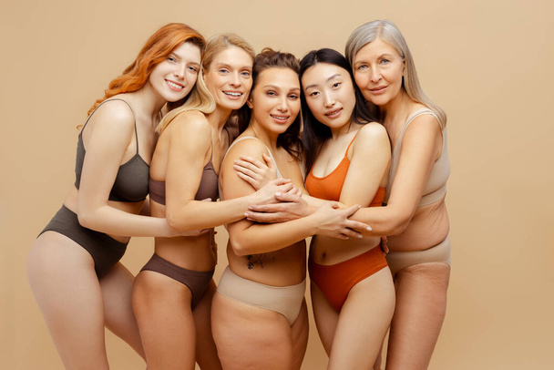 Beautiful smiling diverse multiethnic women wearing stylish sexy lingerie hugging looking at camera, fashion models posing isolated on beige background. International women's day concept - Photo, Image