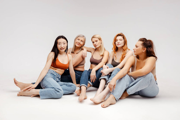 Groep van diverse, lachende vrouwen met stijlvolle jeans en tops, zittend, zittend in de studio. Aantrekkelijke vrouwelijke modellen poseren op witte achtergrond, kijkend naar camera. Begrip diversiteit, reclame - Foto, afbeelding